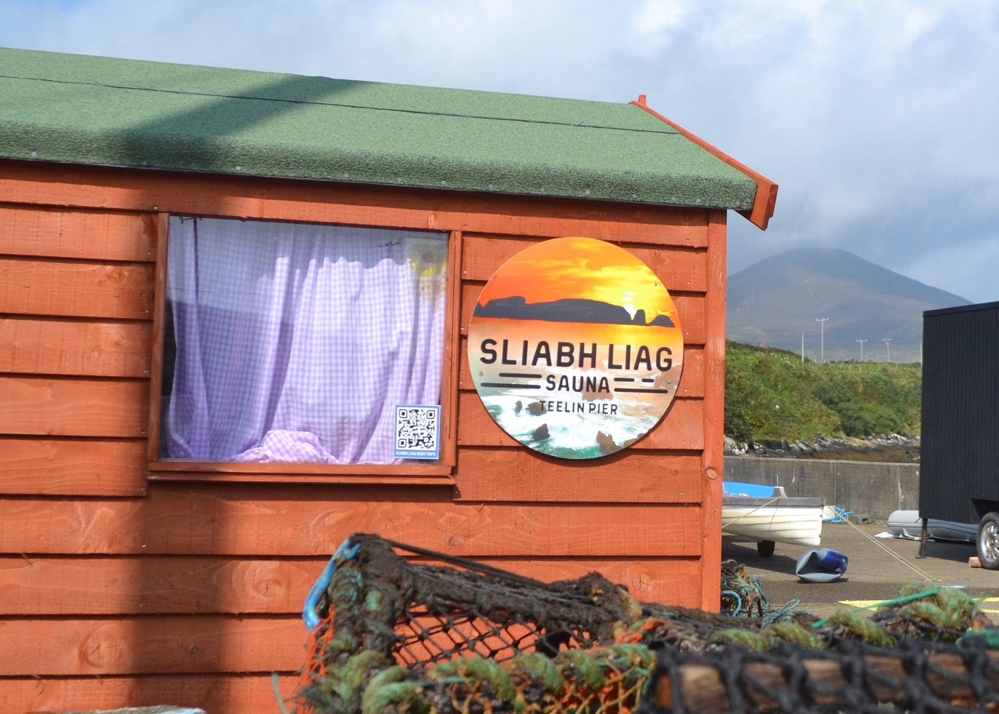 Sliabh Liag Sauna at Teelin Pier in Donegal, with the bespoke double wood-fired sauna beside traditional fishing huts — promotional image for the Sliabh Liag Sauna Gift Card.